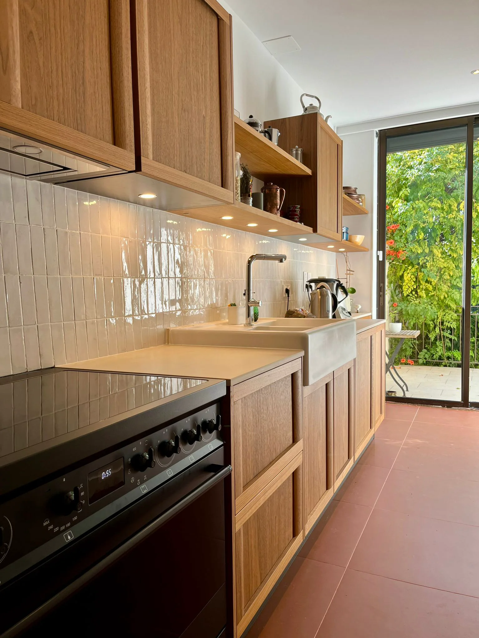 Cuisine étroite avec meubles en bois, évier, four encastré et porte vitrée donnant sur un espace verdoyant.