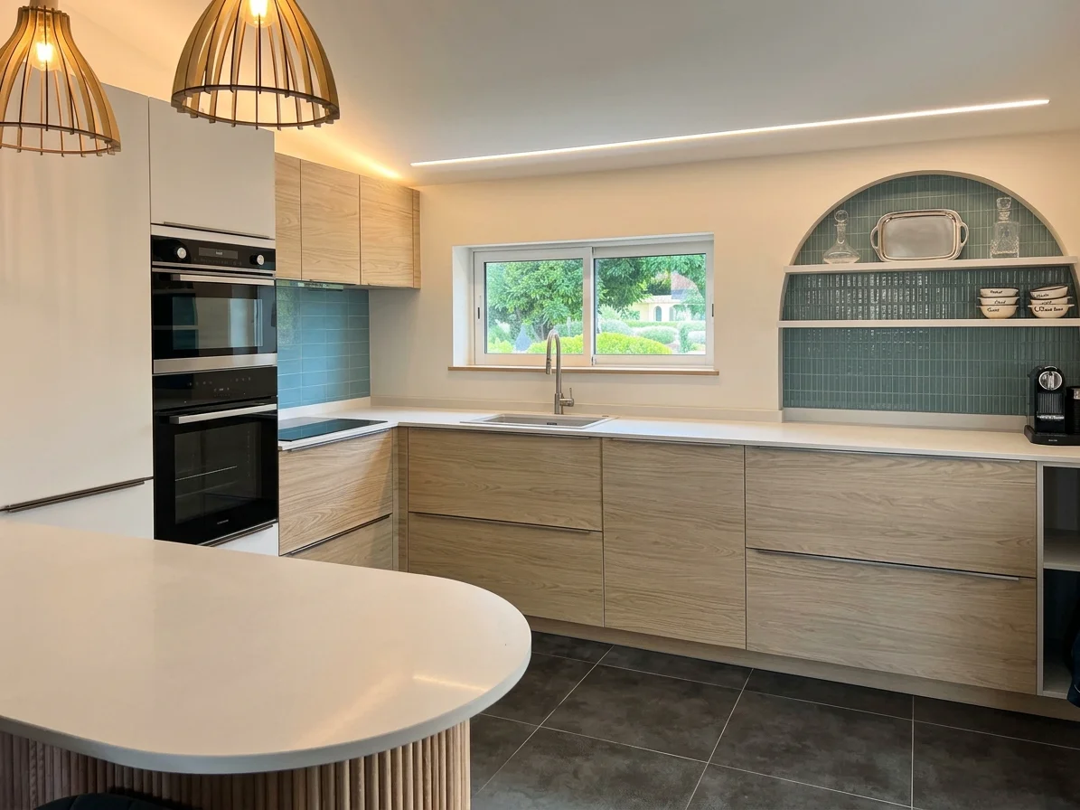 Cuisine équipée en bois avec un long plan de travail blanc, un four encastré, un évier sous une fenêtre et une niche carrelée en arche.