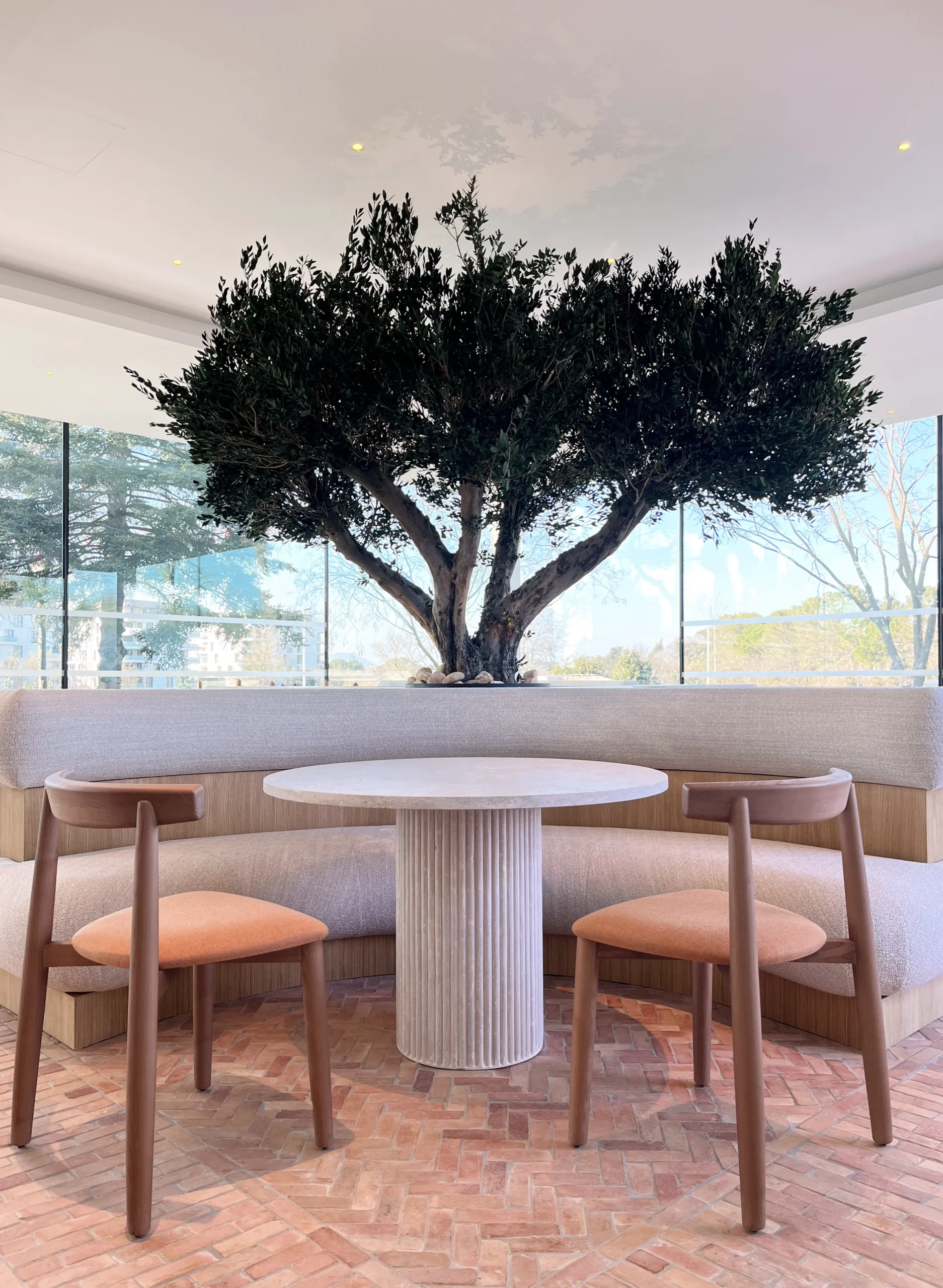 Coin repas vitré avec banquette courbe, table ronde claire et deux chaises en bois à coussin rouille.