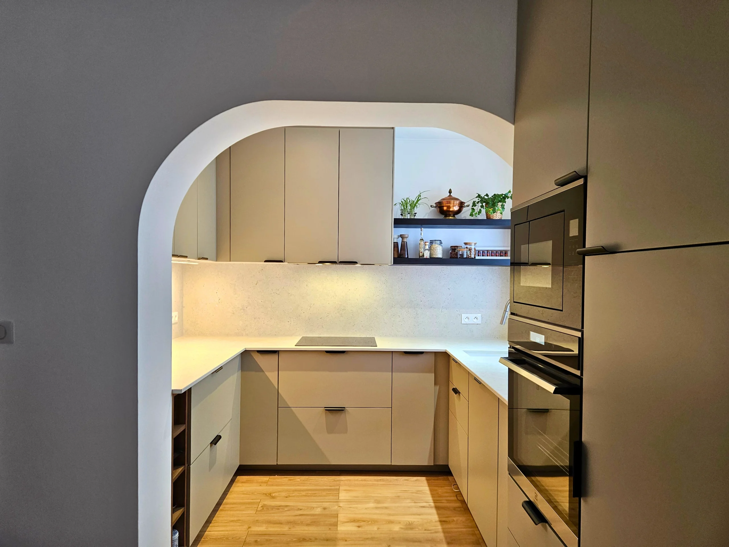 Cuisine compacte en L vue à travers une ouverture arrondie, avec rangements, plan de travail, fours encastrés et sol en bois.
