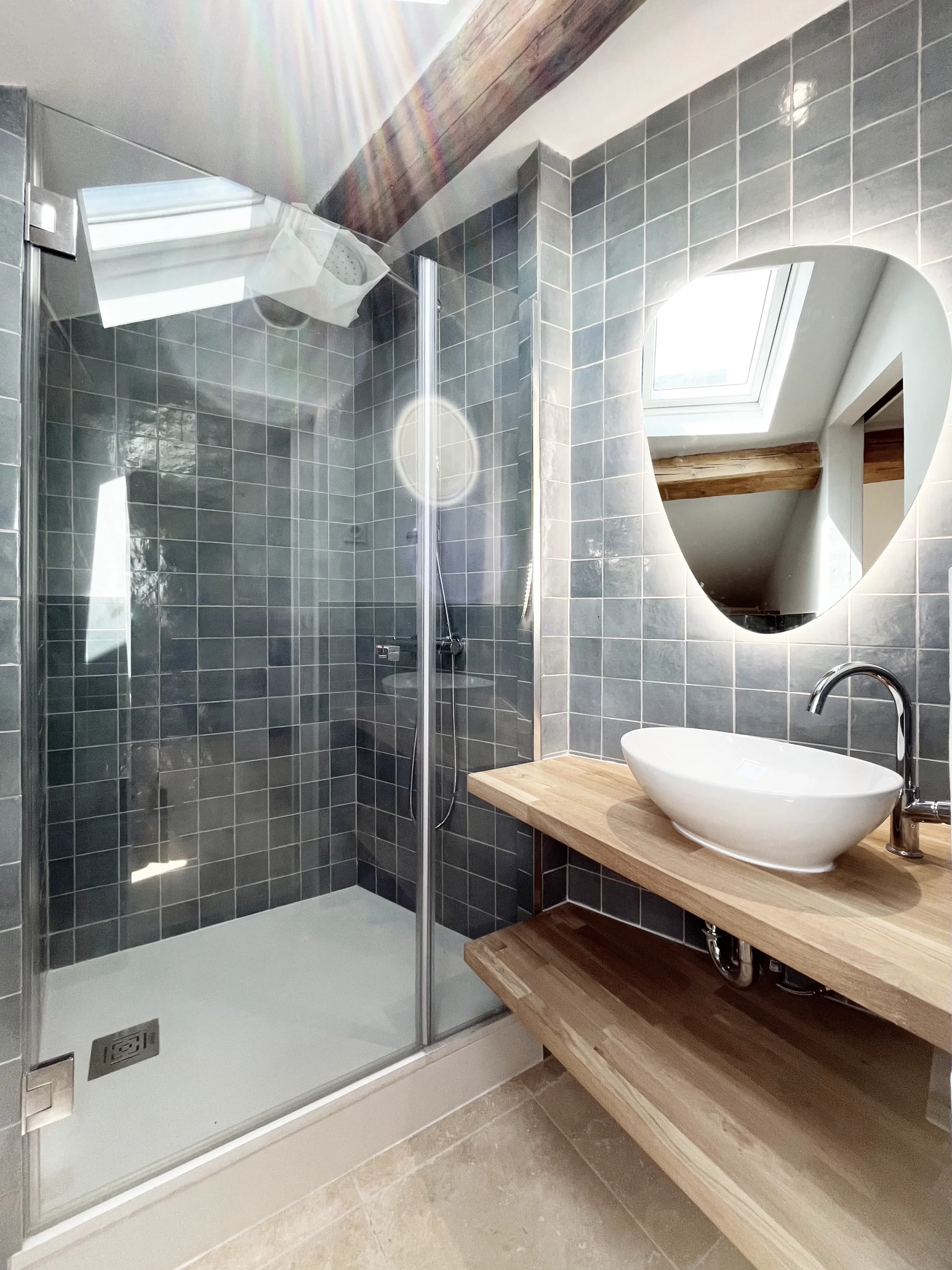 Salle de bain carrelée avec douche vitrée, meuble vasque en bois et lumière naturelle venant d’une fenêtre de toit.