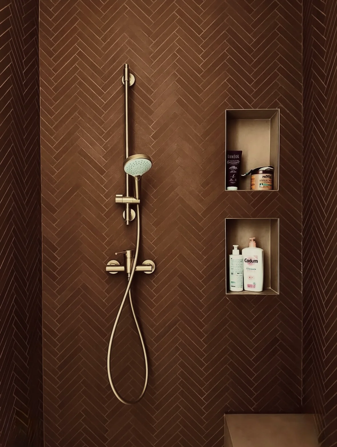 Douche avec carrelage brun posé en chevrons et niches murales contenant des flacons.