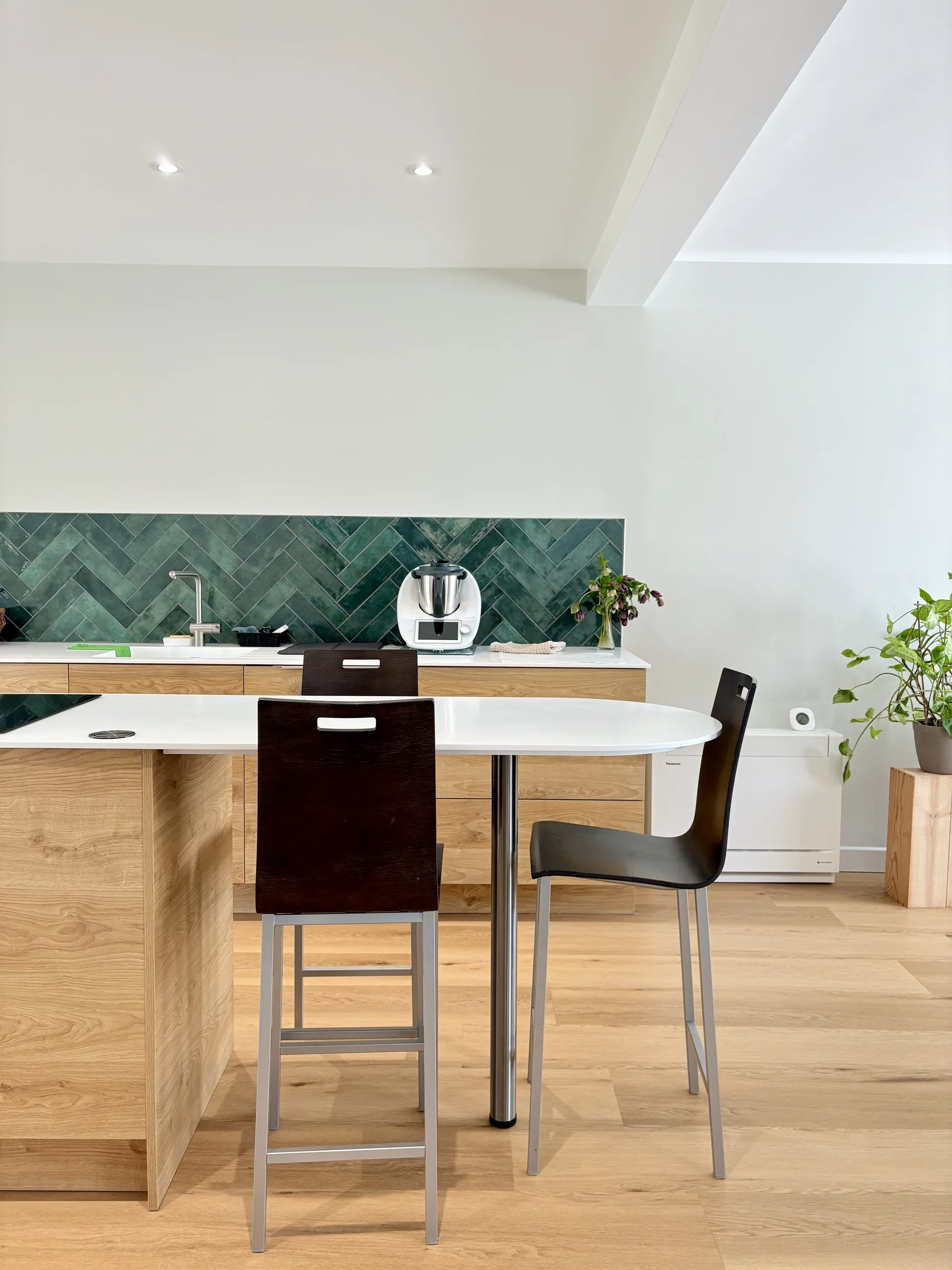 Cuisine et coin repas lumineux avec table blanche, chaises, meubles en bois clair et crédence géométrique verte.