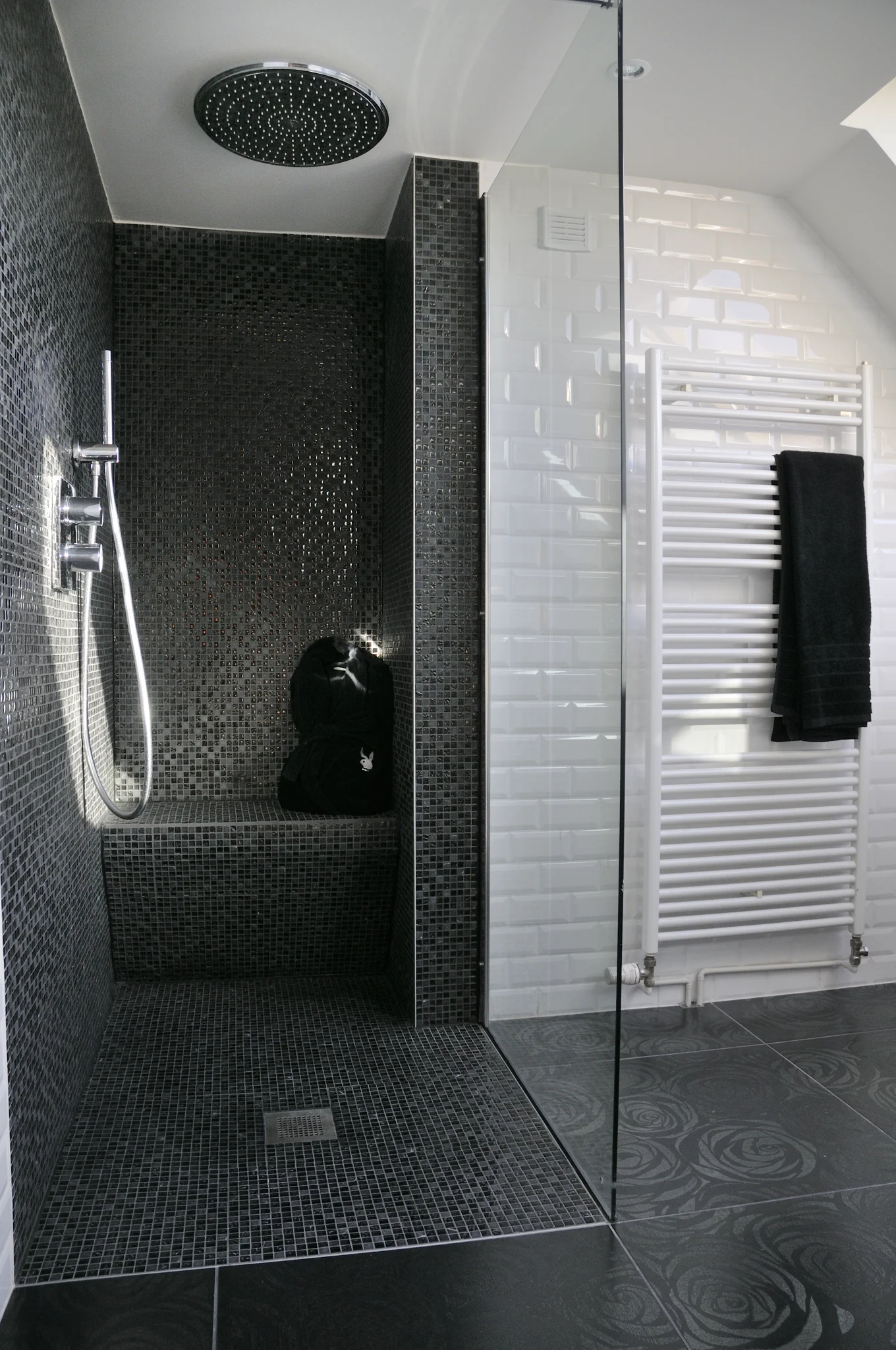 Douche à l’italienne avec paroi vitrée, mosaïque sombre, pomme de douche circulaire et douchette.