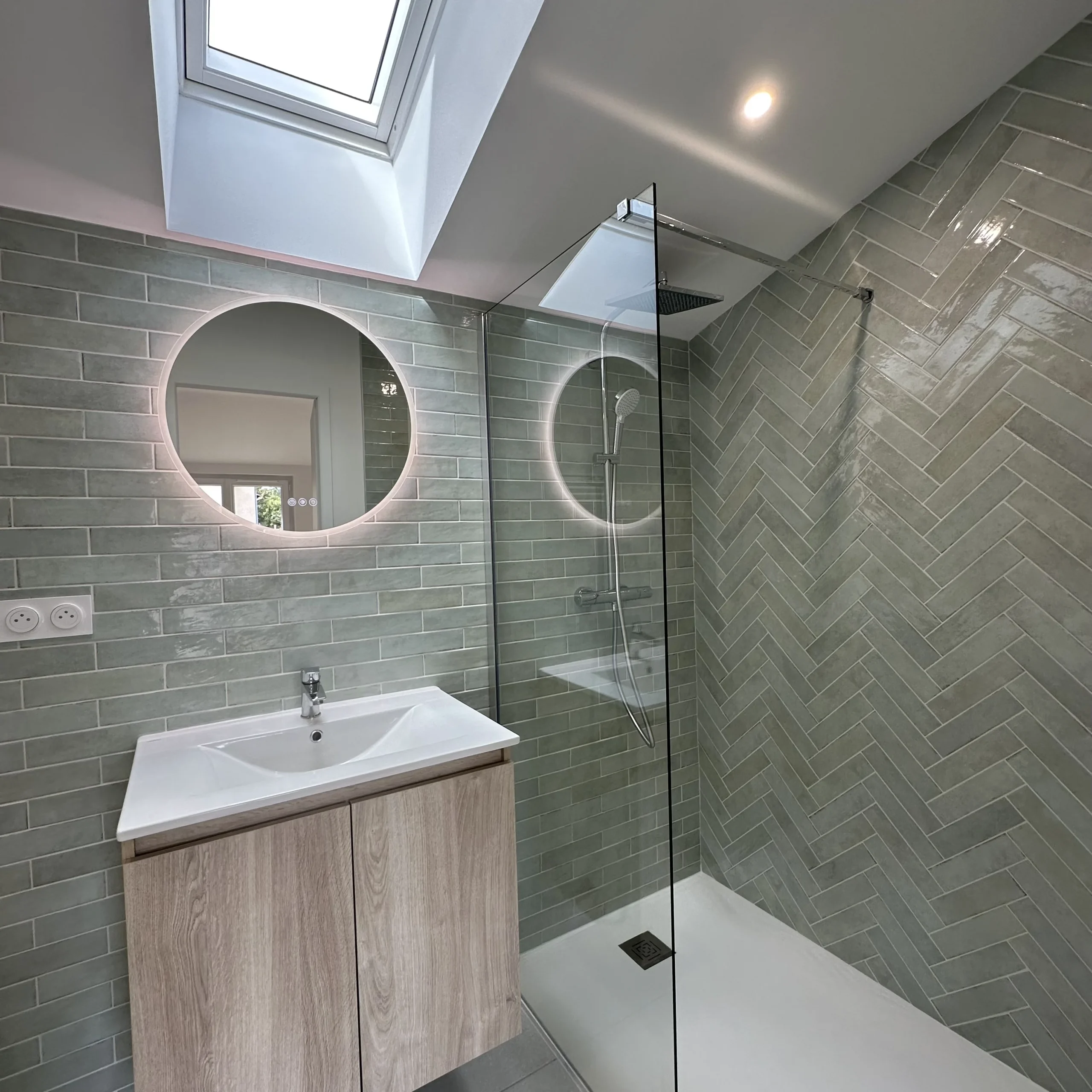 Salle de bain avec douche à l’italienne vitrée, vasque murale, meuble en bois et miroir rond.