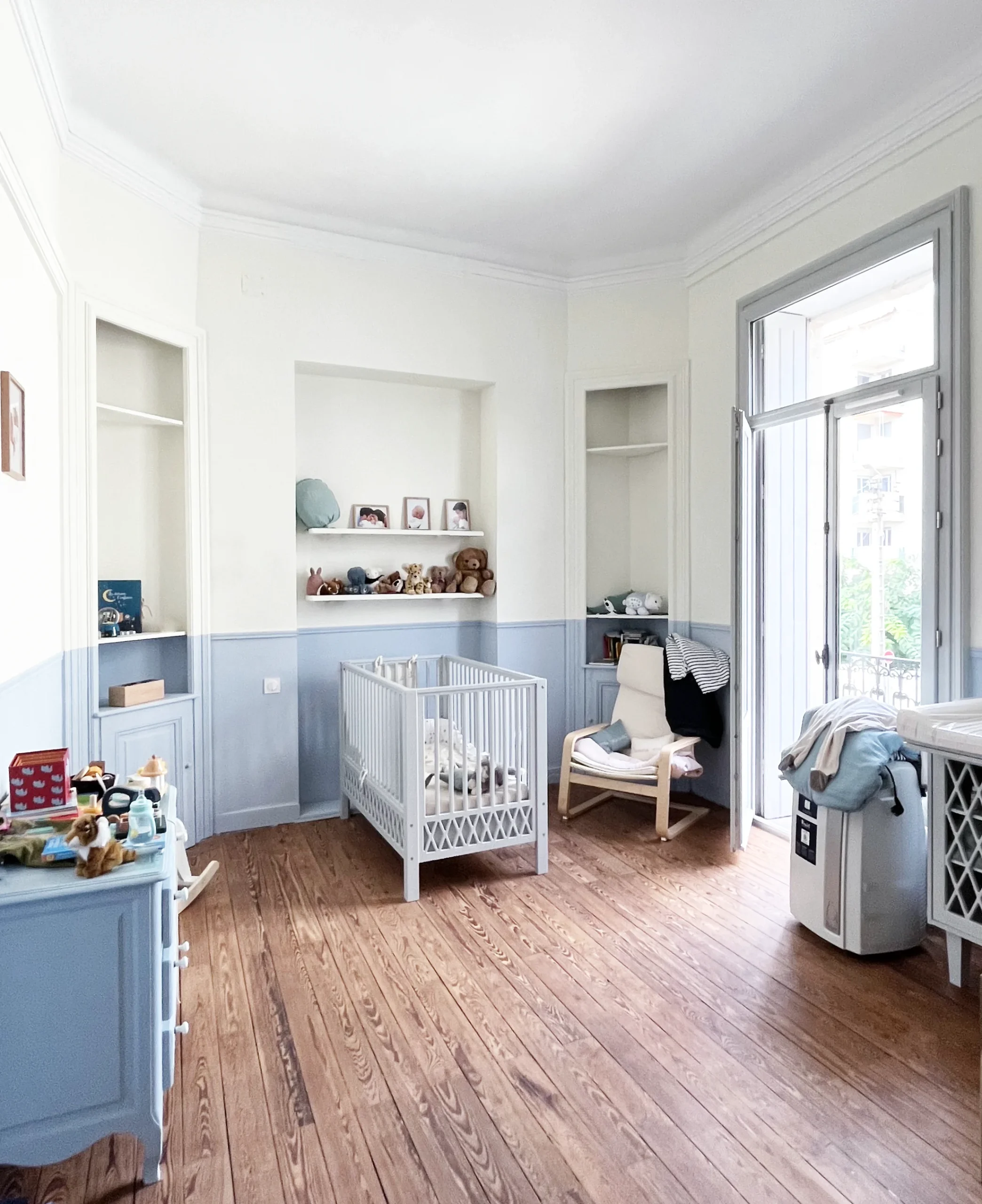 Chambre de bébé lumineuse avec lit à barreaux, étagères intégrées, fauteuil et table à langer près d’une fenêtre.