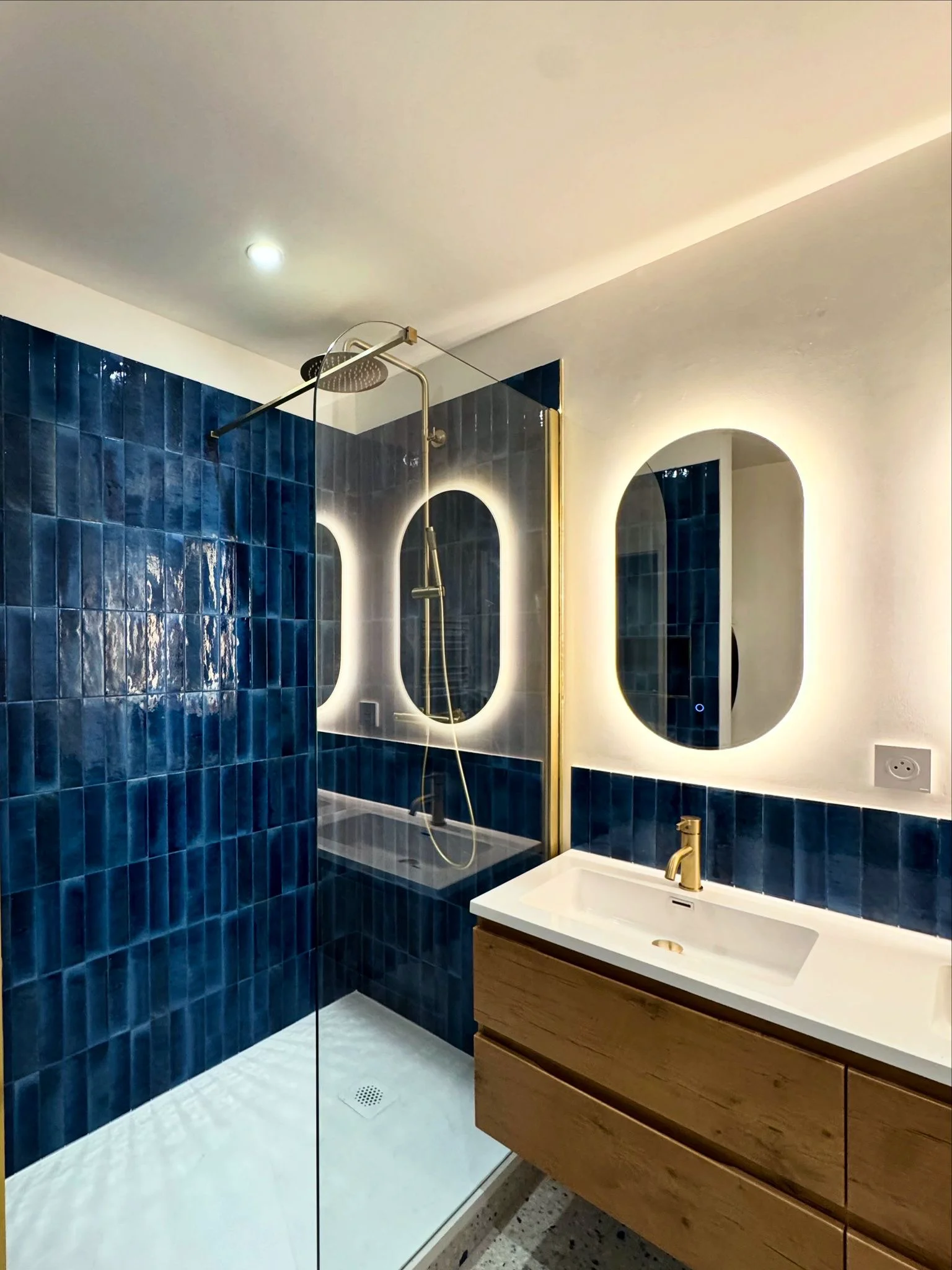 Salle de bain avec douche vitrée, carrelage bleu et meuble vasque en bois.