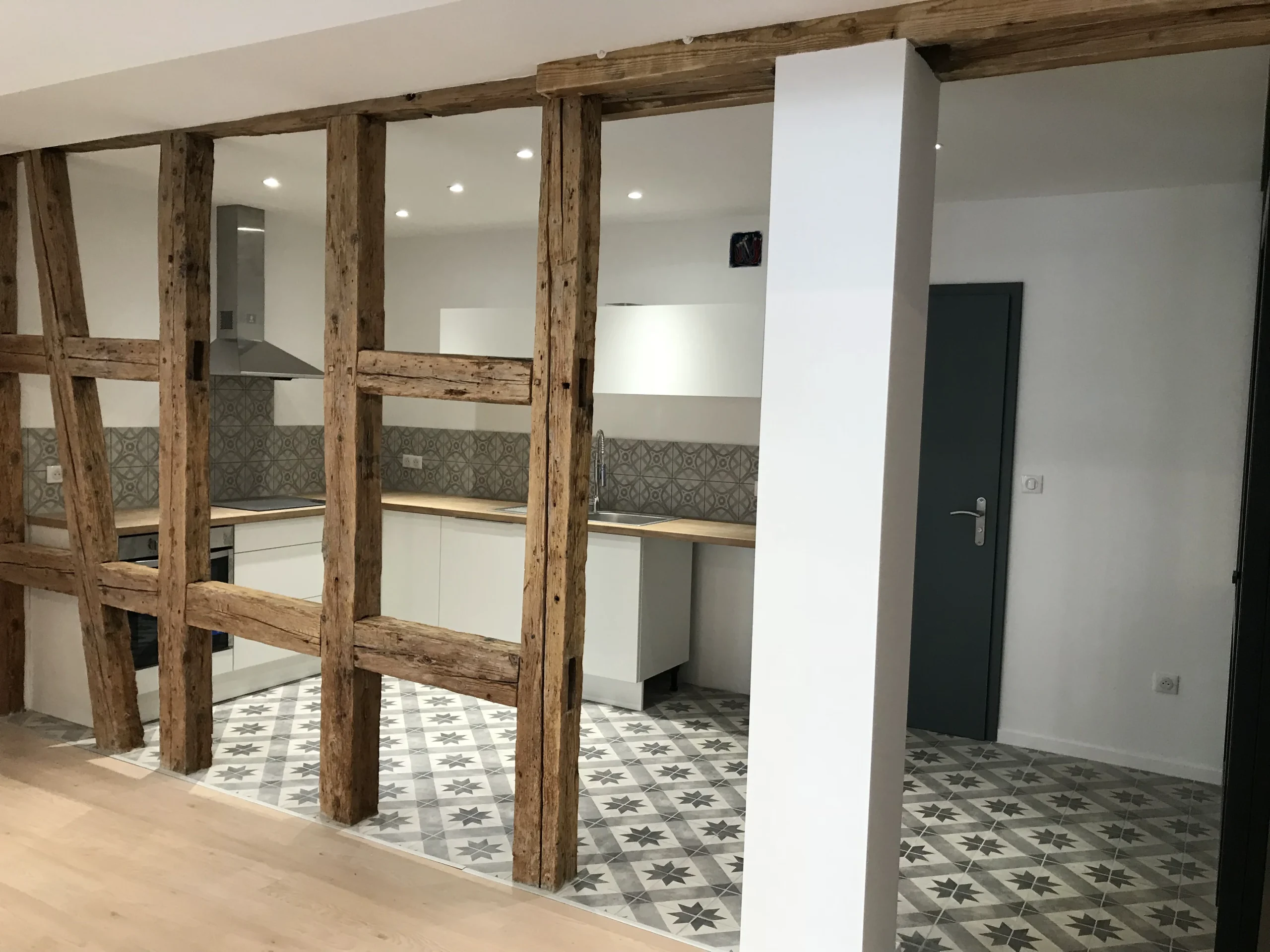 Cuisine en rénovation derrière une ossature en bois apparent, avec des meubles blancs et du carrelage à motifs.